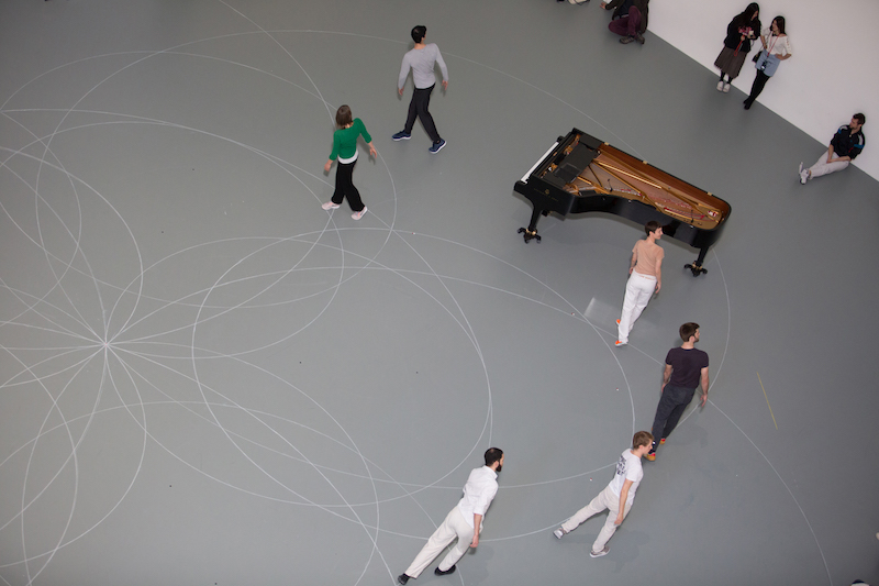 An aerial view of a group of dancers standing on one of the outlines of a circle. They all turn their heads over their shoulders. The piano is also set by them.